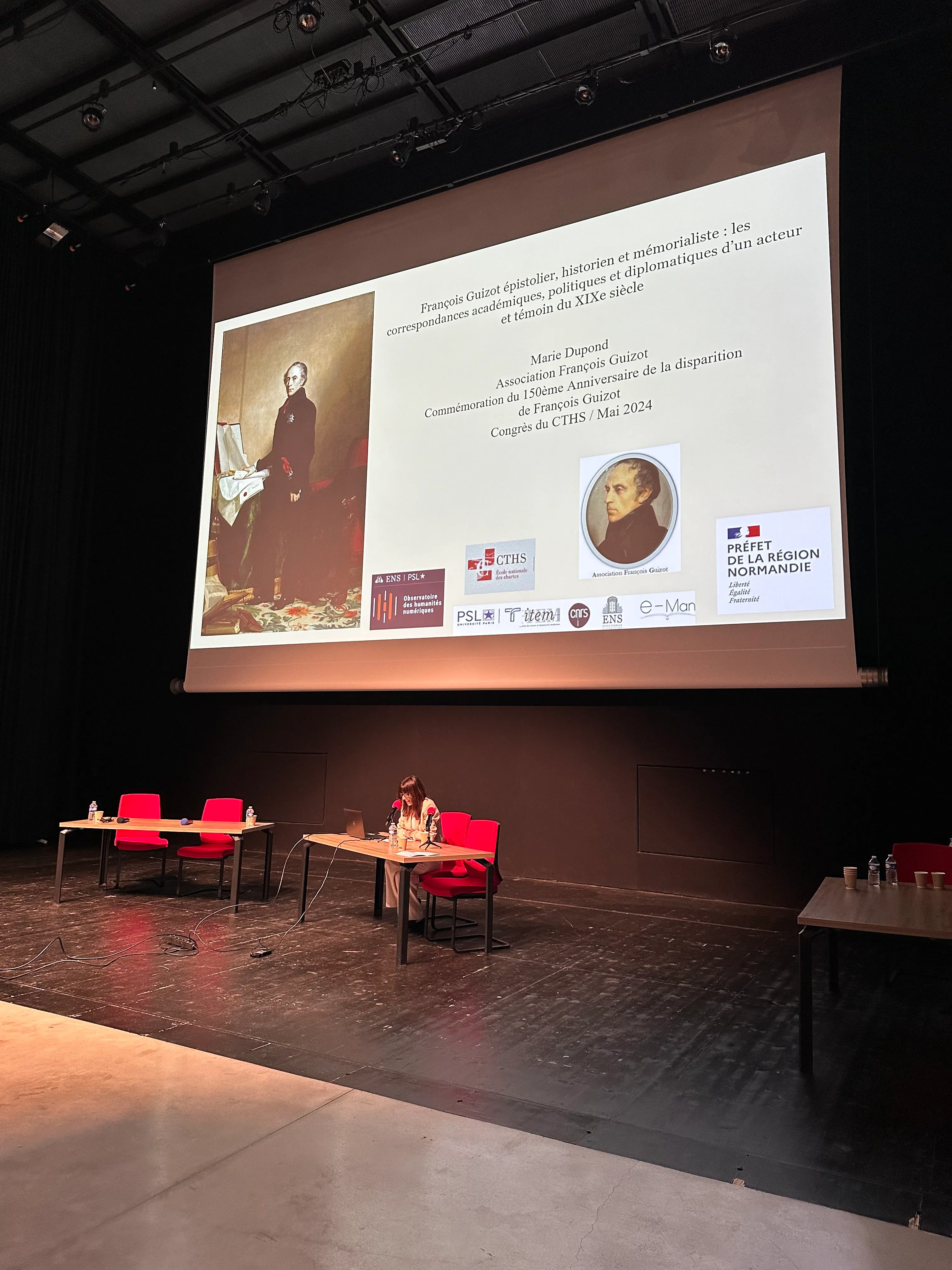 Marie qui fait une conférence avec un écran géant derrière. Elle est assise à un bureau