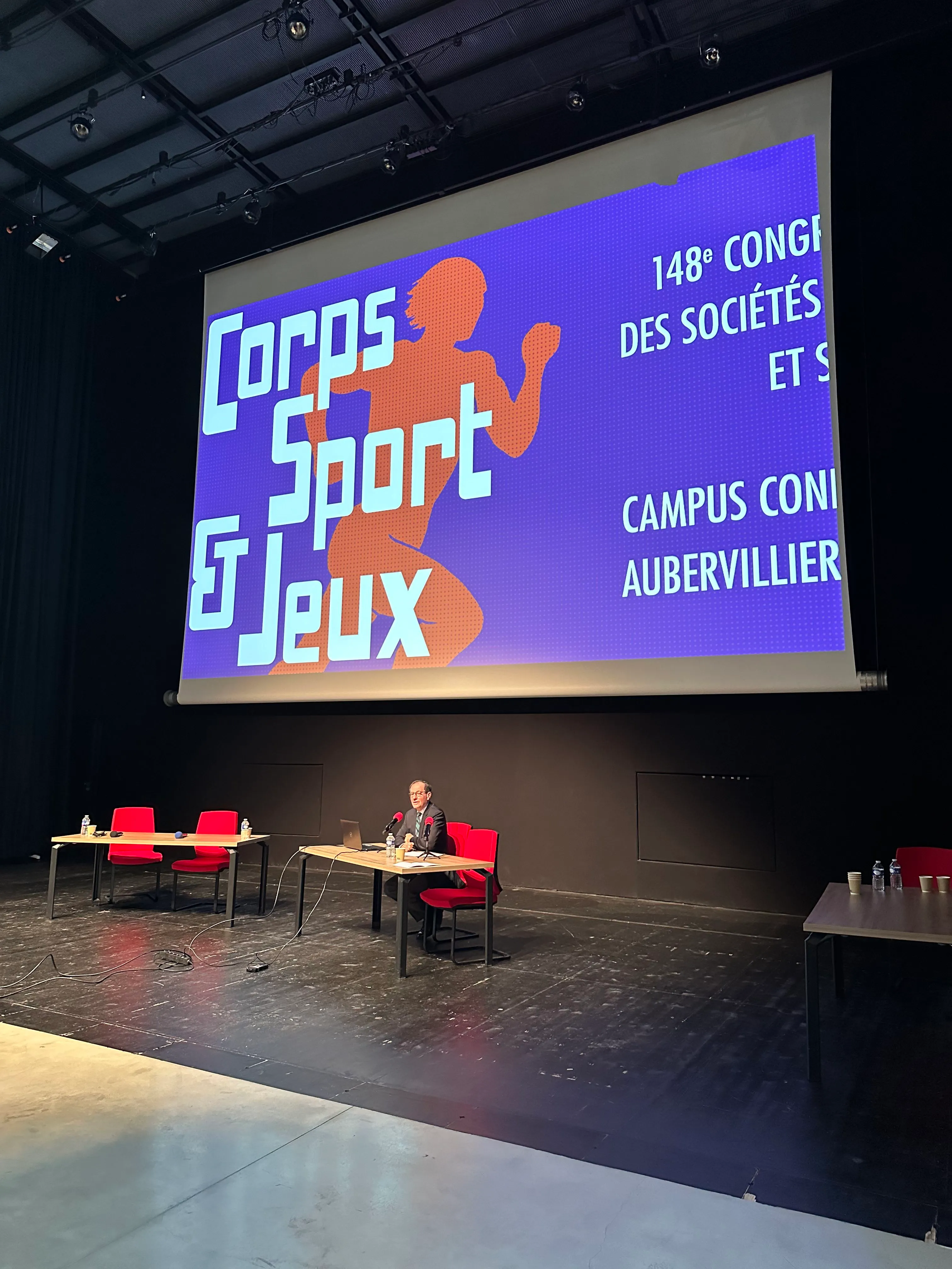 photo du logo corps sport et jeux sur grand écran avec un monsieur assis devant un bureau.
