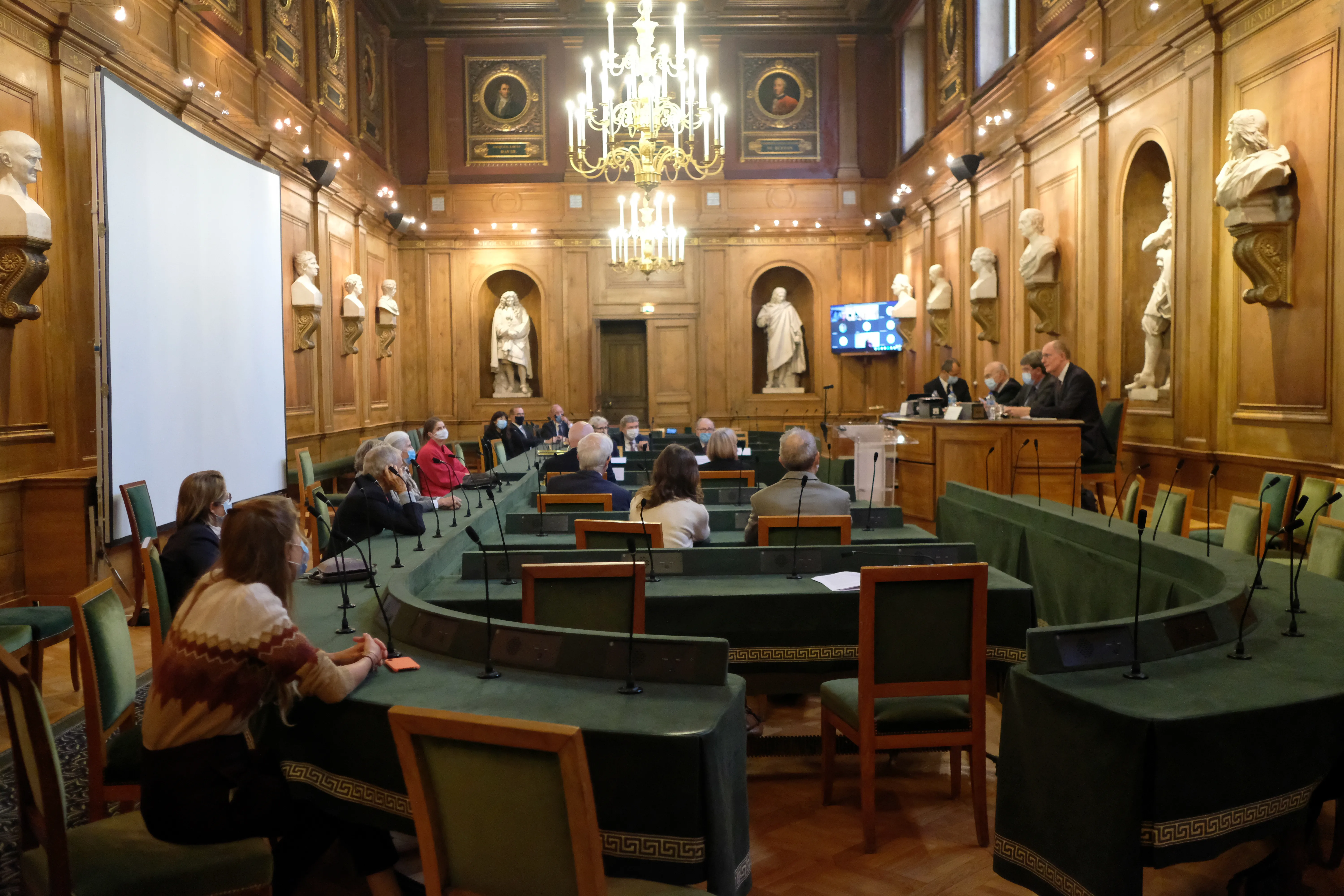 grand pièce de réunion avec des tables vertes, des personnes sont assises et forment un ovale. Elles ont des micros disposés devant elles.